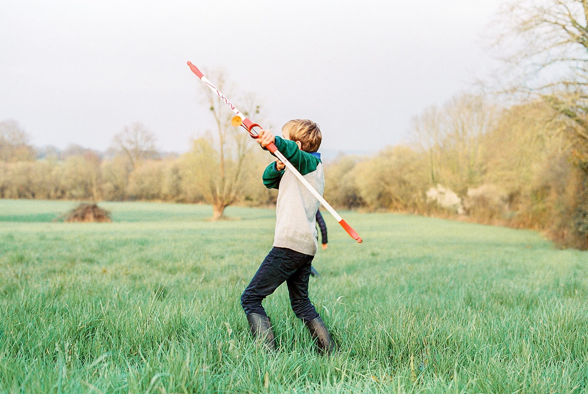 A young boy plays with a rubber bow and arrow set in a large grassy field at sunset. 