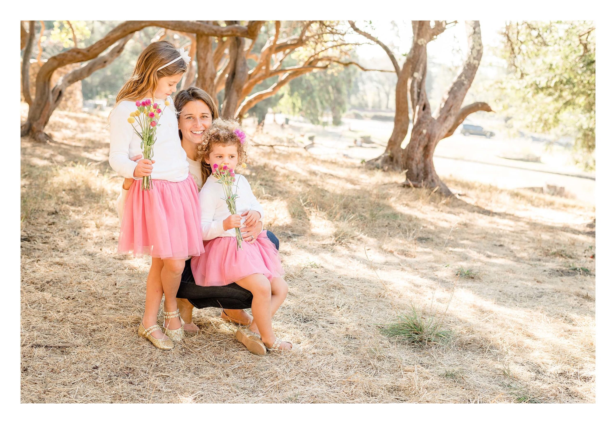A mother kneels on the ground, balancing her younger daughter on one knee while the older one stands next to her. The girls hold pink and yellow bouquets of flowers and wear fluffy pink tutus.