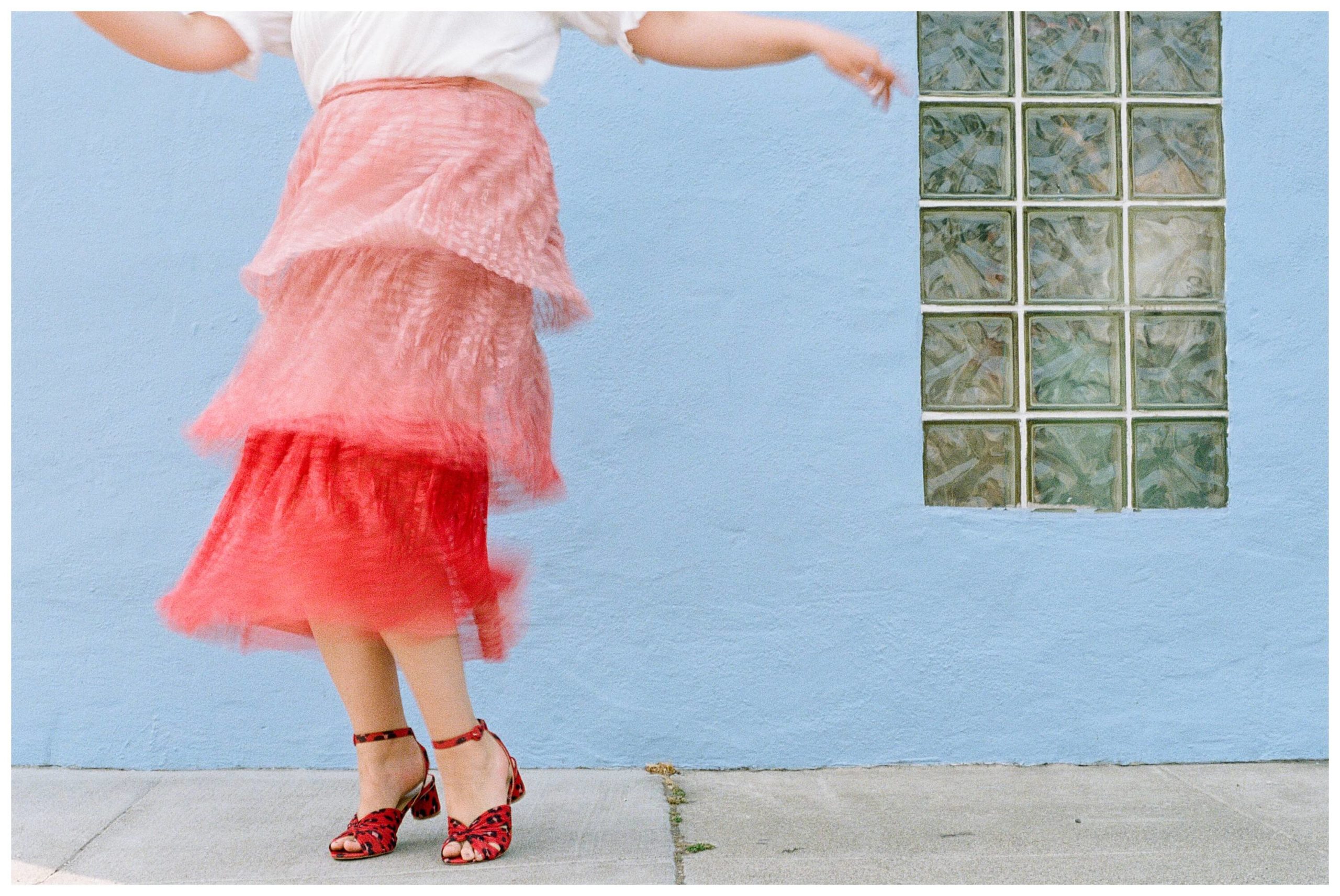 A girl wearing a white shirt and ombré red skirt with red heels twists and turns in front of a blue wall. You can see the movement of her skirt as she dances.