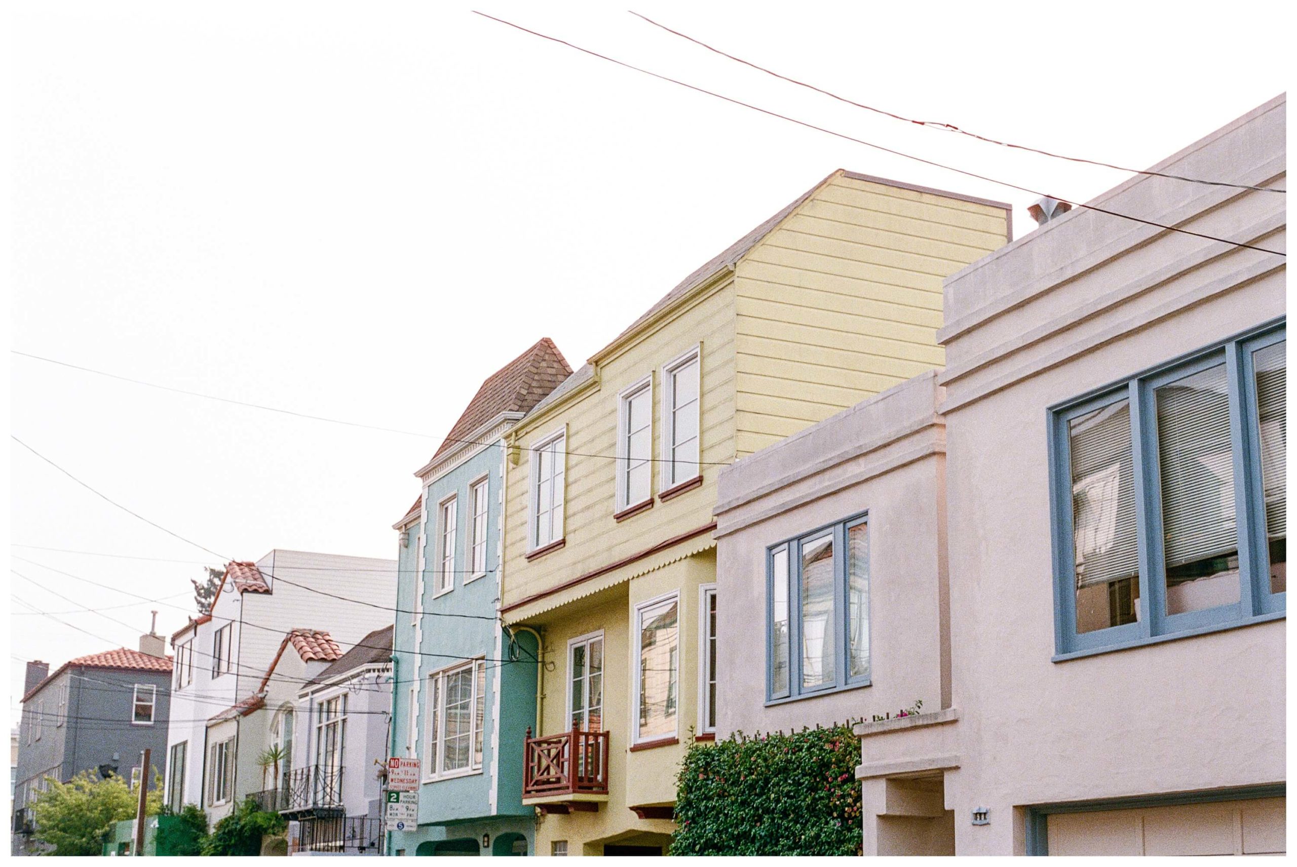 A row of colorful houses in shades or pastel pink, yellow, and blue line the street at sunset in the Mission District of San Francisco.