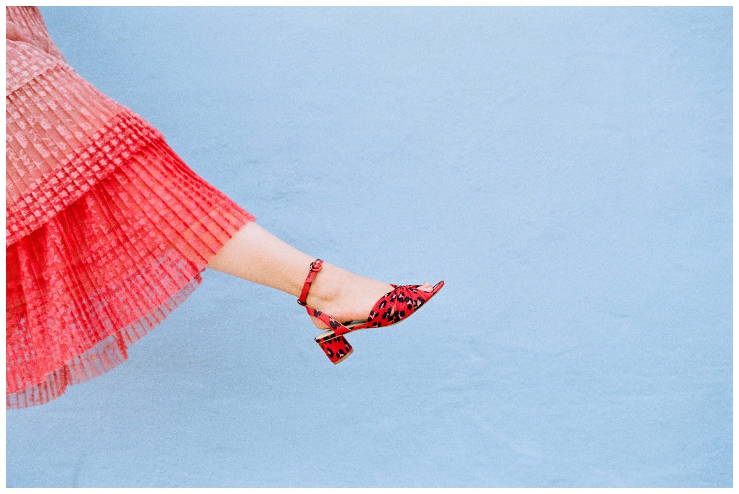 A girl playfully kicks up one leg in front a blue wall, the obmbré red of her skirt and the deep red of her high heels add a playful element. 
