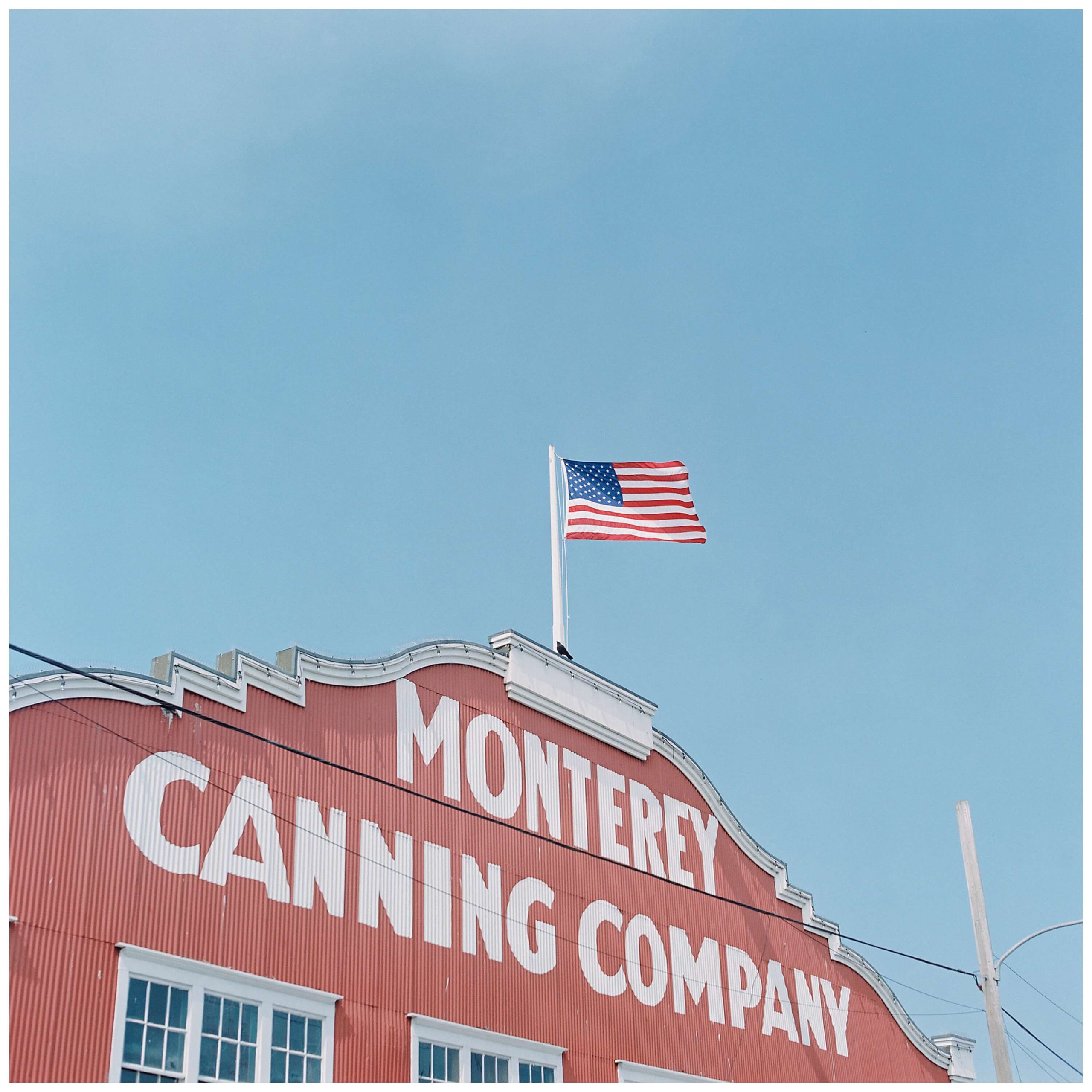 The iconic red building on Cannery Row in Monterey reads "Monterey Canning Company" in large, capital white letters. An American flag sits centered on the roof against a blue sky.