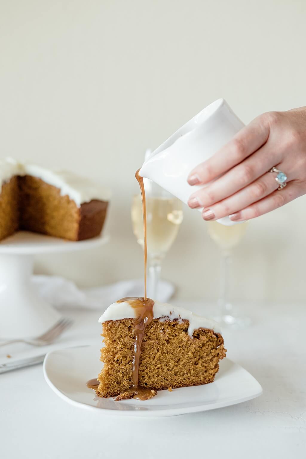 Coconut sugar caramel sauce is drizzled on top of a triangular slice of iced pumpkin cake. The cake sits on a pedestal in the background next to two filled champagne flutes.