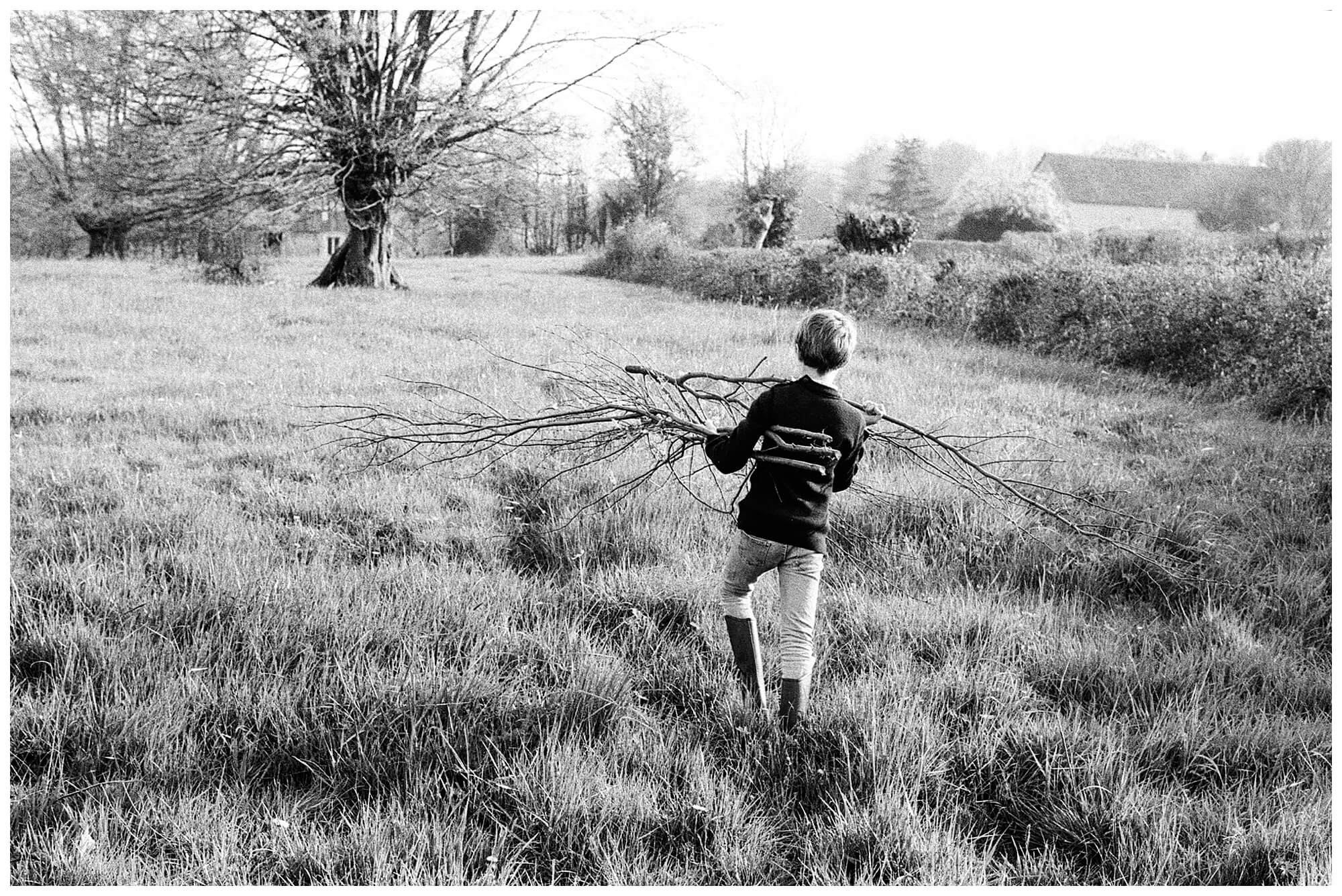 A young boy hauls two large tree branches through a grassy field in the French countryside.