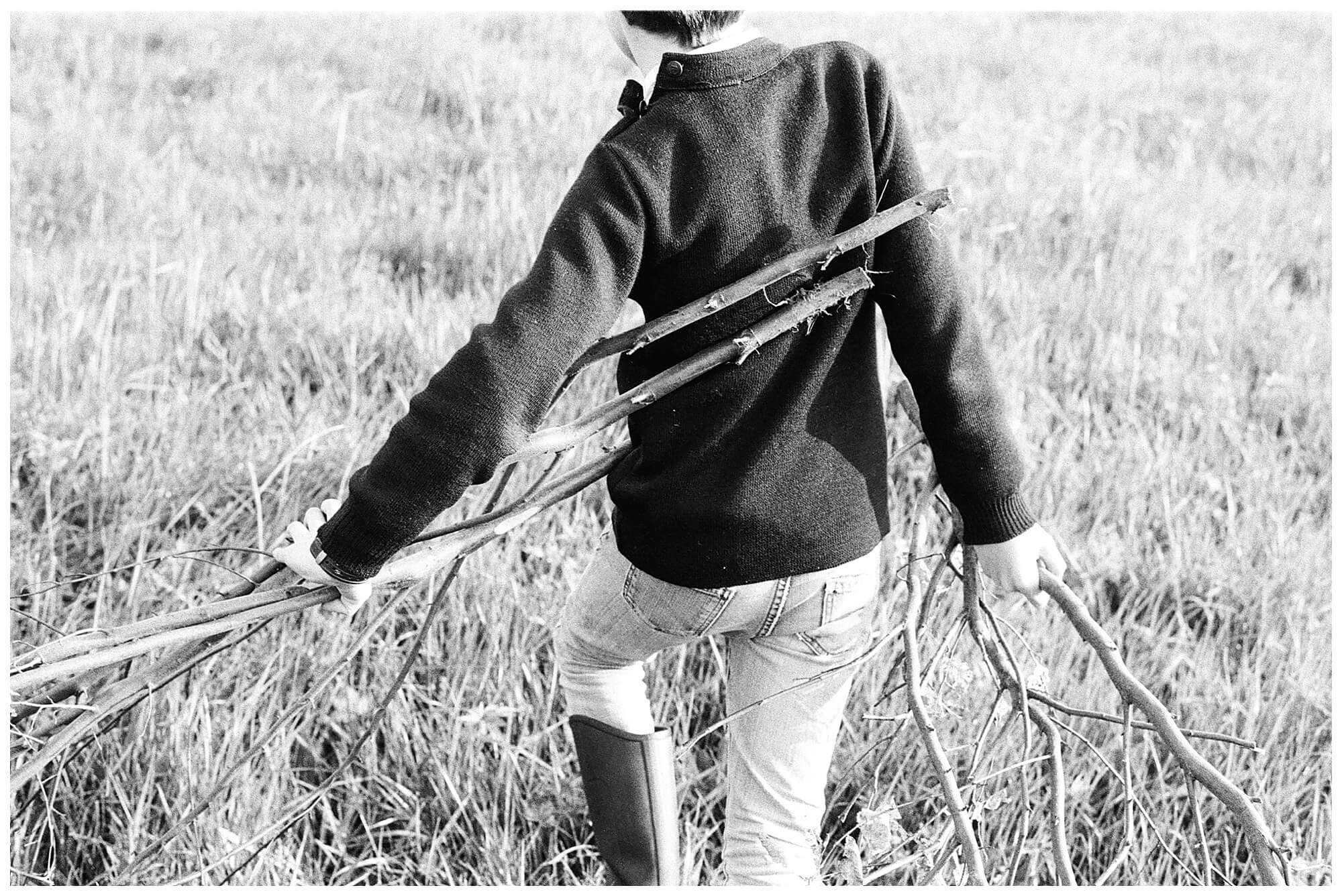 A young boy carries two large tree branches through a grassy field in the French countryside. 
