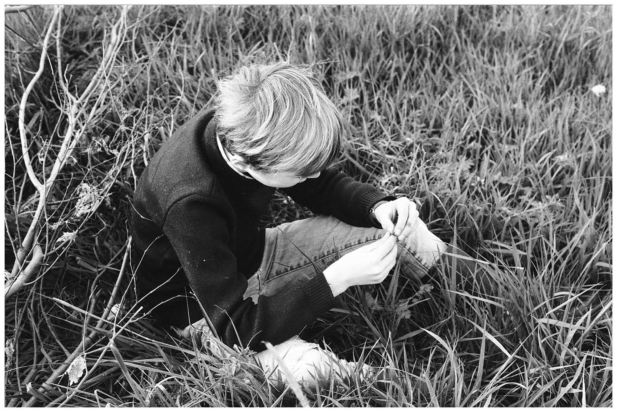 Black and white image of a young boy sitting amongst the tall grasses in a field in the French countryside.