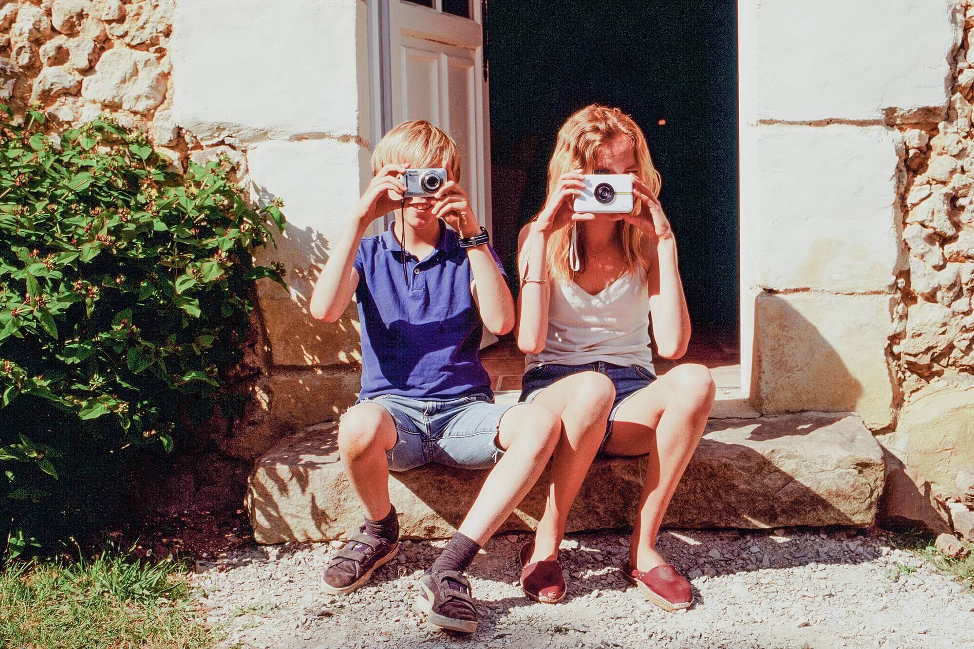 Two children, a boy on the left and girl on the right, sit on the stoop of a stone French country cottage during the COVID 19 quarantine. They are holding up cameras in front of their faces.