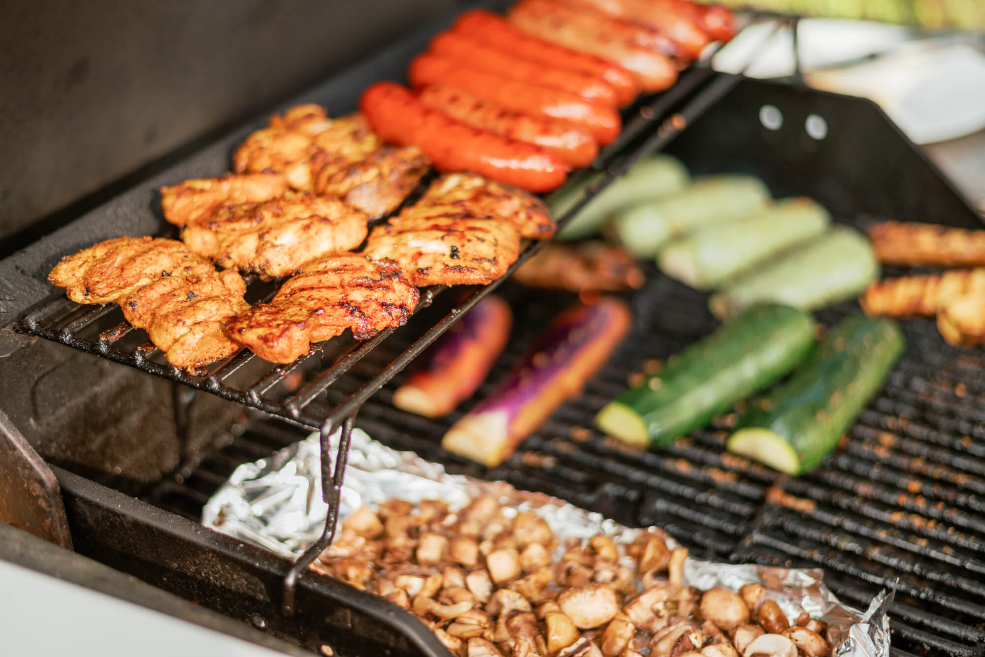 Chicken, sausages, and vegetables sit on the grill.