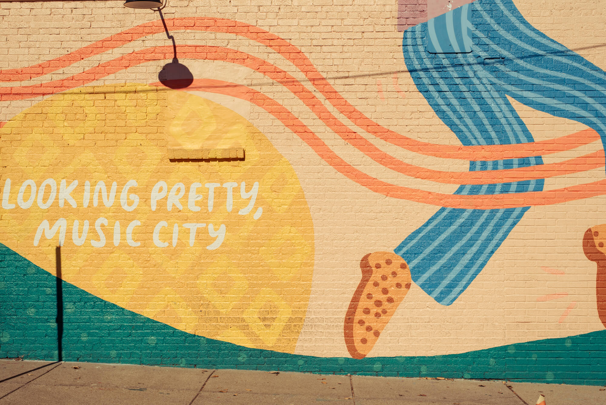 large green, orange, and yellow mural on a brick wall in Nashville's 12 South neighborhood. It features a pair of dancing legs wearing striped blue pants and polk-a-dot orange and red shoes. The white hand-written text says "Looking pretty Music City"